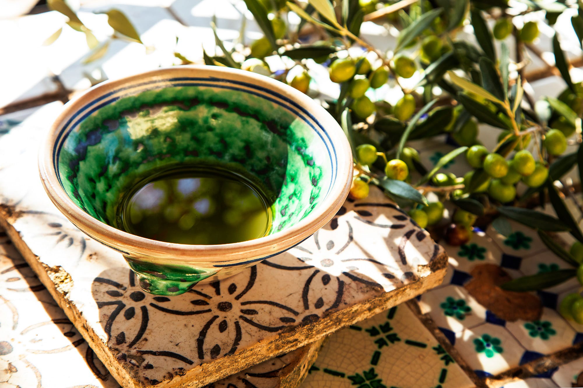 Grøn keramisk skål med frisk olivenolie omgivet af olivengrene og modne oliven